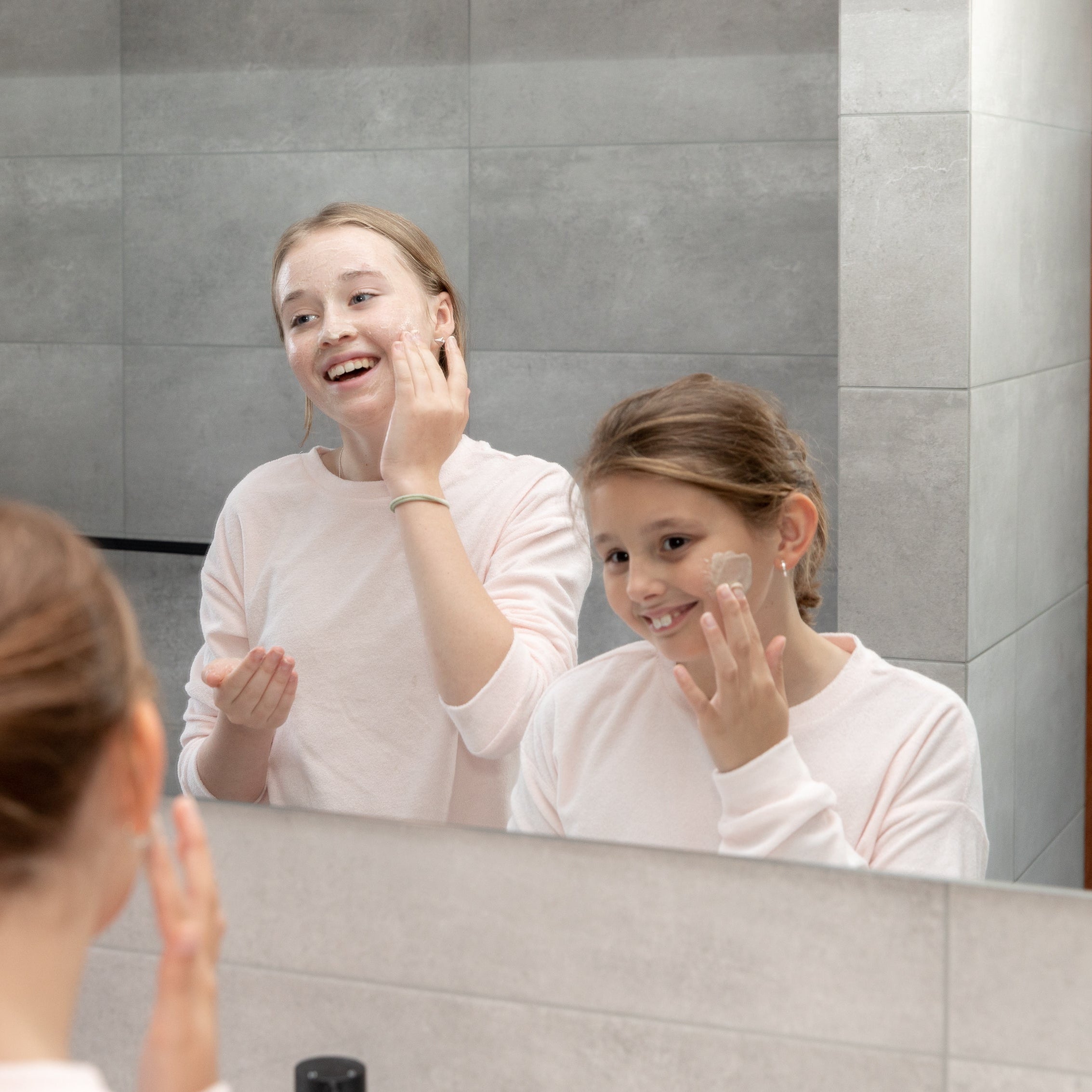 Two teens applying cream to their faces in front of a bathroom mirror.